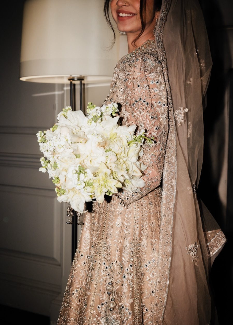 Bride in an embellished champagne gown holding a white bouquet of stock and roses in soft lamplight